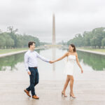 A couple holds hands and walks past the Reflecting Pool with the Washington Monument in the background