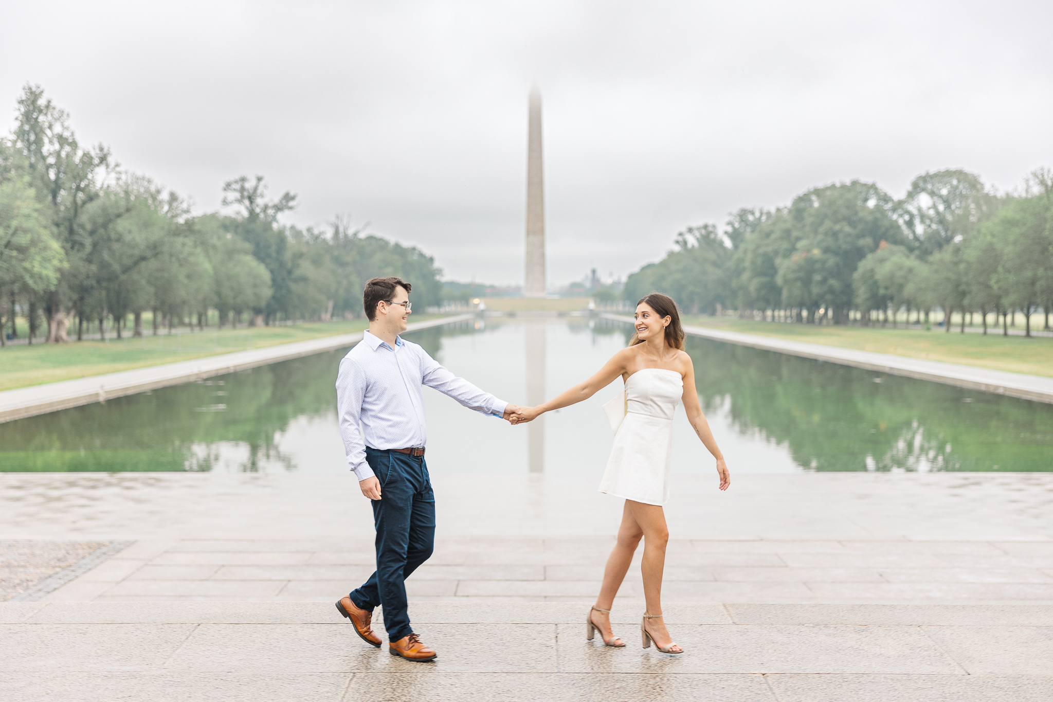 A couple holds hands and walks past the Reflecting Pool with the Washington Monument in the background